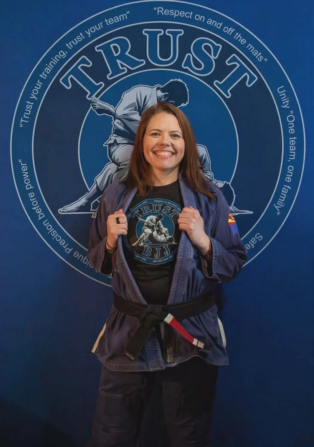 BJJ instructor Missy Baize in a navy gi and black belt, smiling in front of the Trust BJJ academy logo wall.